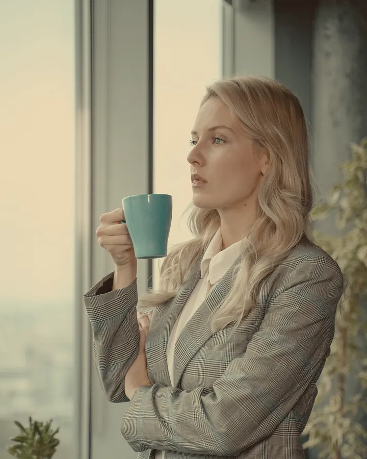 A woman in a grey checked blazer holding a coffee mug and looking out of an office window