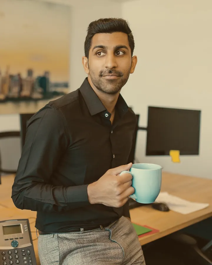 A man in a black shirt holding a coffee mug, leaning against a desk in an open-plan office