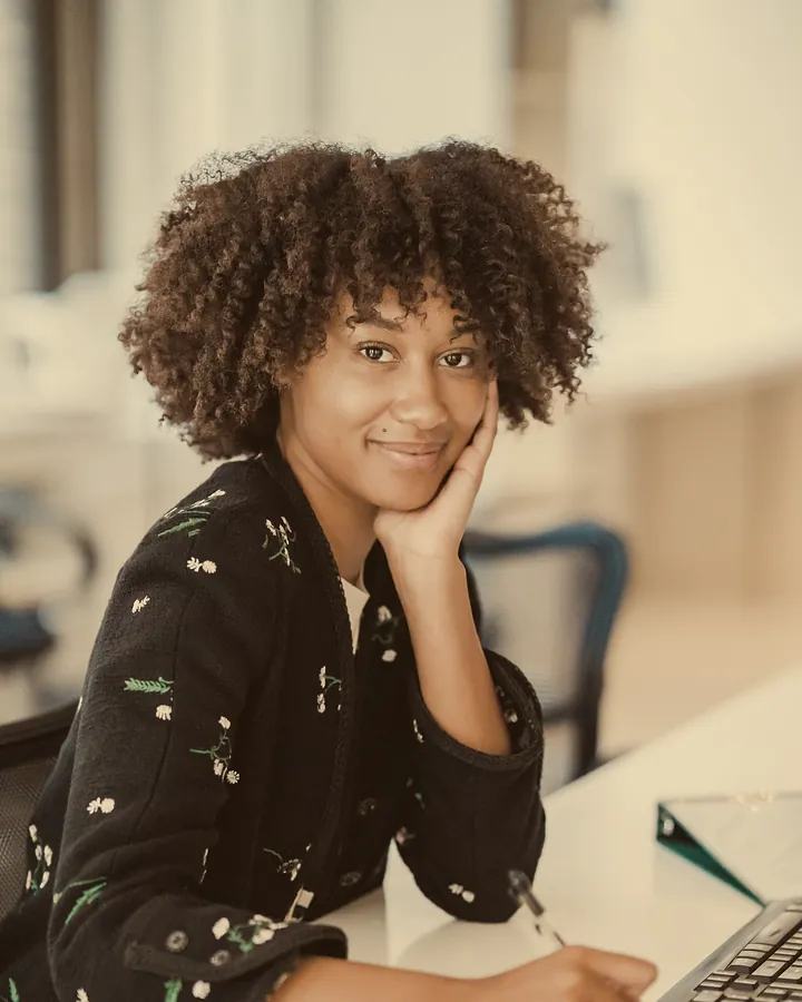 A smiling woman with curly hair sitting at an office desk, resting her chin on her hand